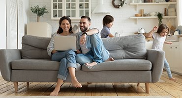 Couple sitting on living room couch looking at laptop while two children play tag behind them.