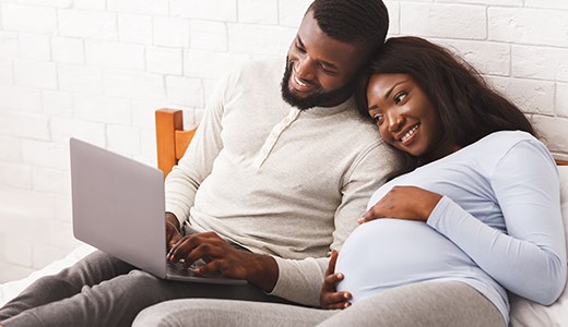 Expectant couple reclining on made bed looking at plan benefits on laptop computer